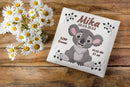 Geschenkidee zur Geburt mit Koalabär auf Kissen – niedlicher Koala mit rosa Ohren und offenem Mund, dekoriert mit stilvollen Angaben zur Geburt.