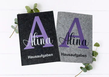 Zwei Filzhüllen für Hausaufgabenhefte mit großem lila Anfangsbuchstaben auf grauem und schwarzem Hintergrund.