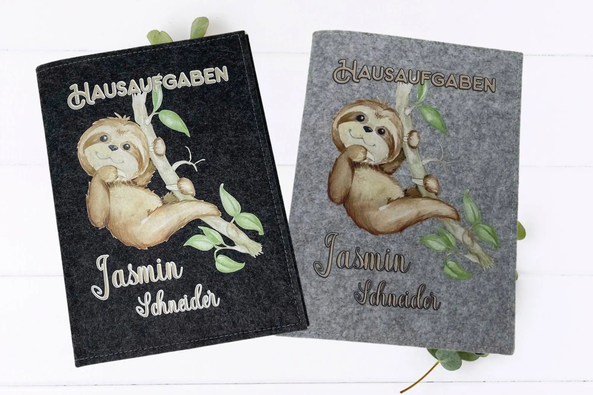 Schulheft-Hüllen mit Faultier-Design, das an einem Ast hängt, auf grauem Filzhintergrund.
