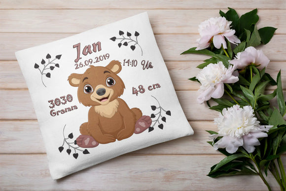 Verspieltes Bärenkissen als Geburtsgeschenk – Mittelpunkt bildet ein fröhlicher kleiner Bär, umgeben von stilisierten Schriftzügen mit Geburtsdaten.
