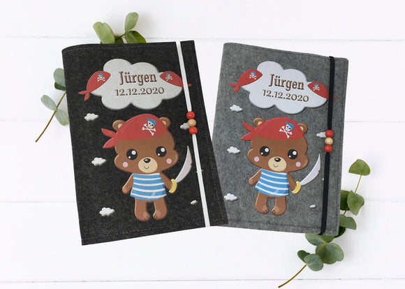 Personalisierte U-Heft-Hülle aus grauem Filz mit Piratenbär, rotem Kopftuch und Wolkenmotiv.
