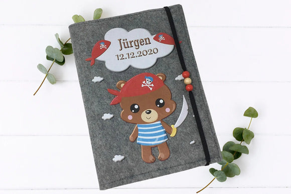 Handgemachte Filzhülle fürs Untersuchungsheft mit Seeräuberbär, Wolkenhimmel und Holzperlenband.