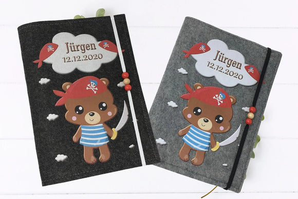 U-Heft-Hülle mit Bärenmotiv als Seeräuber, verziert mit roten und gelben Holzperlen am Gummiband.