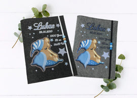 Personalisierte U-Heft-Hülle aus grauem Filz mit schlafendem Teddybär auf blauer Decke und Sternenmütze.