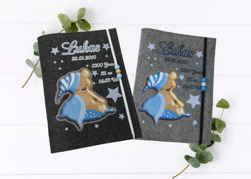 Personalisierte U-Heft-Hülle aus grauem Filz mit schlafendem Teddybär auf blauer Decke und Sternenmütze.