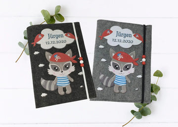 Personalisierte U-Heft-Hülle aus grauem Filz mit süßem Piraten-Waschbär und rotem Kopftuch.