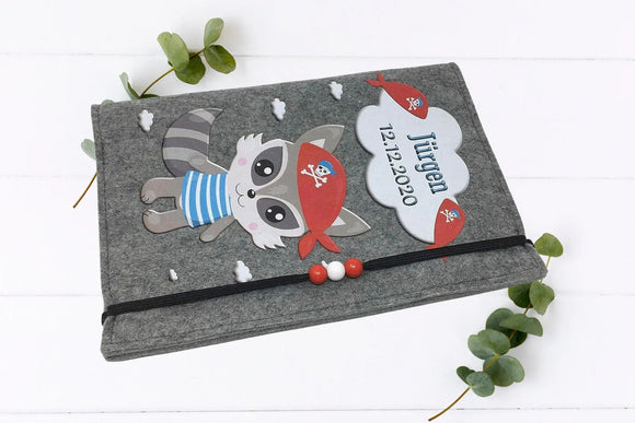 Verspieltes Design mit Piraten-Waschbär und Sprechblase für Namen und Datum auf U-Heft-Hülle.“