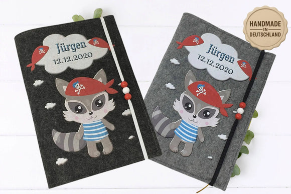 U-Heft-Hülle mit lustigem Waschbär in Seeräuber-Verkleidung, Wolken und bunten Holzperlen.