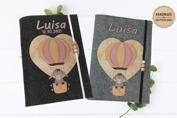 Graue Filzhülle fürs U-Heft mit Eselmotiv im Heißluftballon vor gemustertem Herz-Hintergrund.