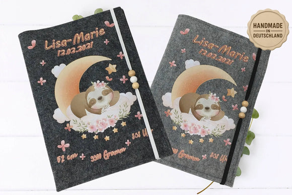 U-Heft-Hülle mit schlafendem Baby-Faultier auf Halbmond, rosa Blüten und Holzkugel-Gummiband.