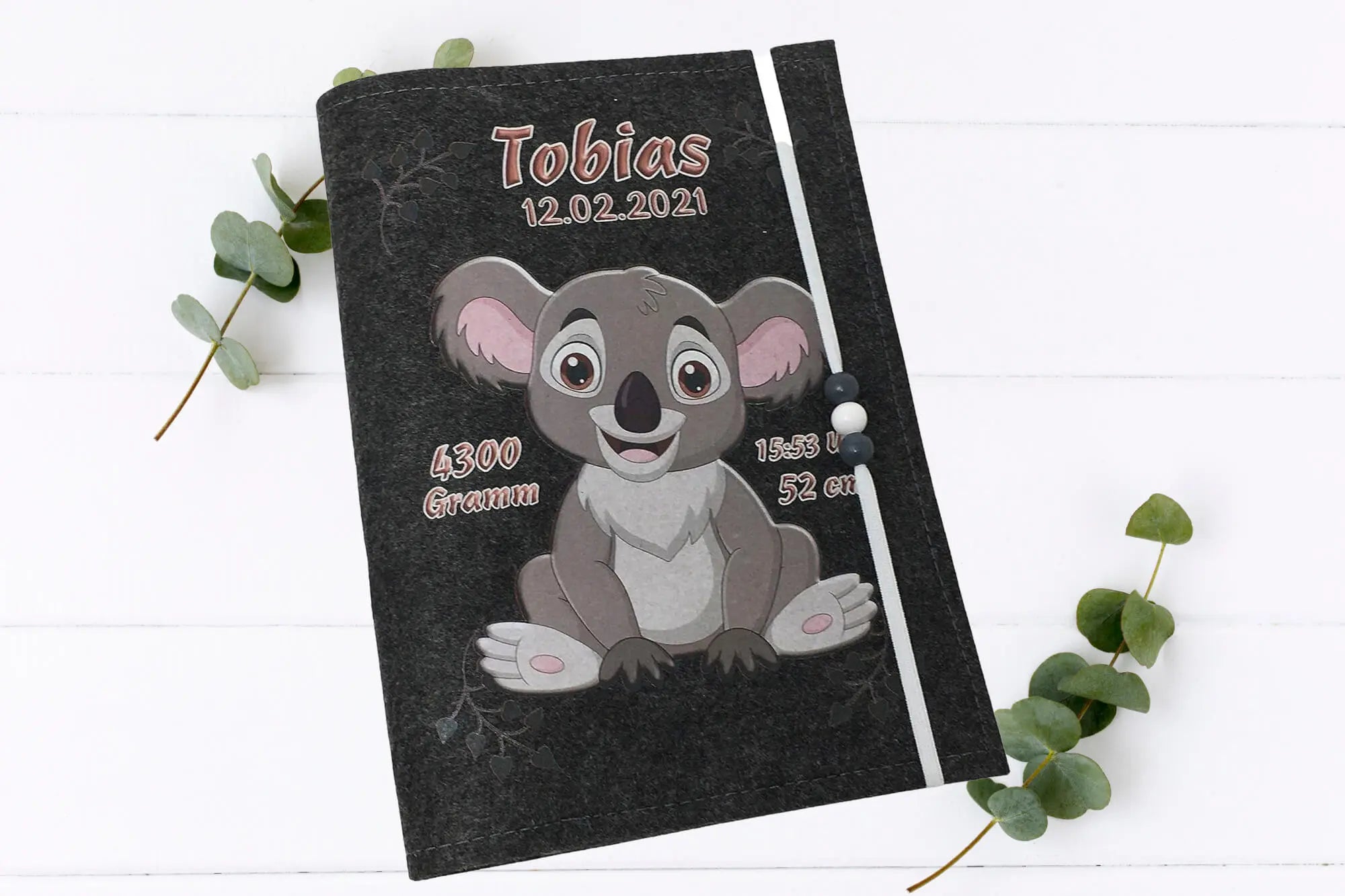 Niedliches Koala-Baby auf U-Heft-Hülle aus Filz – verziert mit schwarzen Herzmustern und rosa Details.