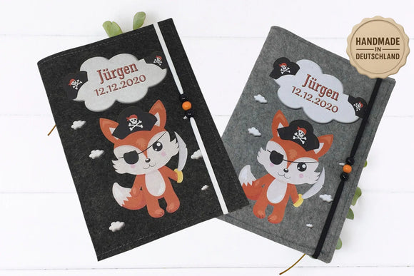 Illustrative U-Heft-Hülle mit Piratenfuchs und Säbel, verziert mit Piratenhüten und Wolken auf grauem Filz.