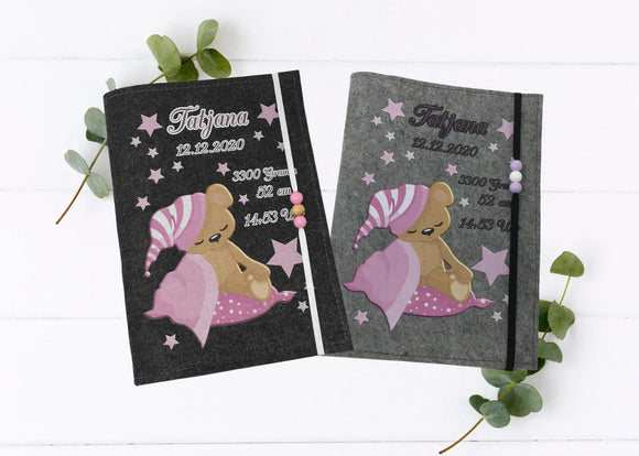 Personalisierte U-Heft-Hülle aus grauem Filz mit schlafendem Teddybär auf rosa Decke und Sternenmütze.