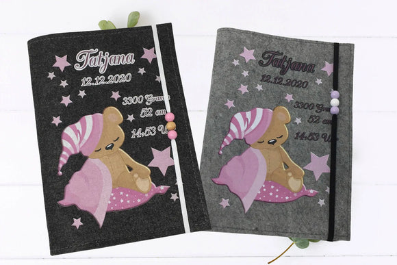 Graue Filzhülle fürs U-Heft mit schlafendem Teddy, rosa Sternenhimmel und elastischem Band mit Perlen.