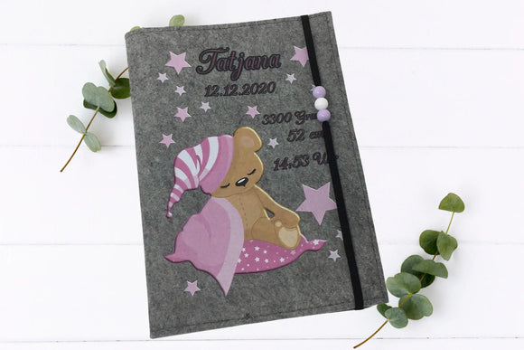 Handgemachte U-Heft-Hülle mit schlafendem Teddy, rosa Nachthimmel und Perlenband zur Fixierung.