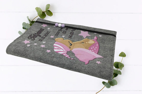 Verspielte U-Heft-Hülle mit schlafendem Bärchen auf Decke, rosa Mütze und funkelnden Sternen.