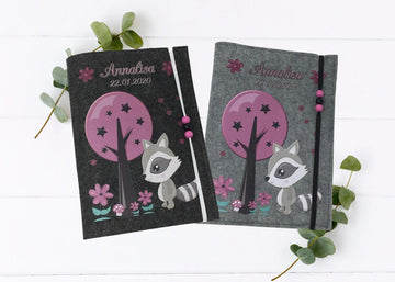 Personalisierte U-Heft-Hülle aus anthrazitfarbenem Filz mit süßem Waschbär und lila Baum.