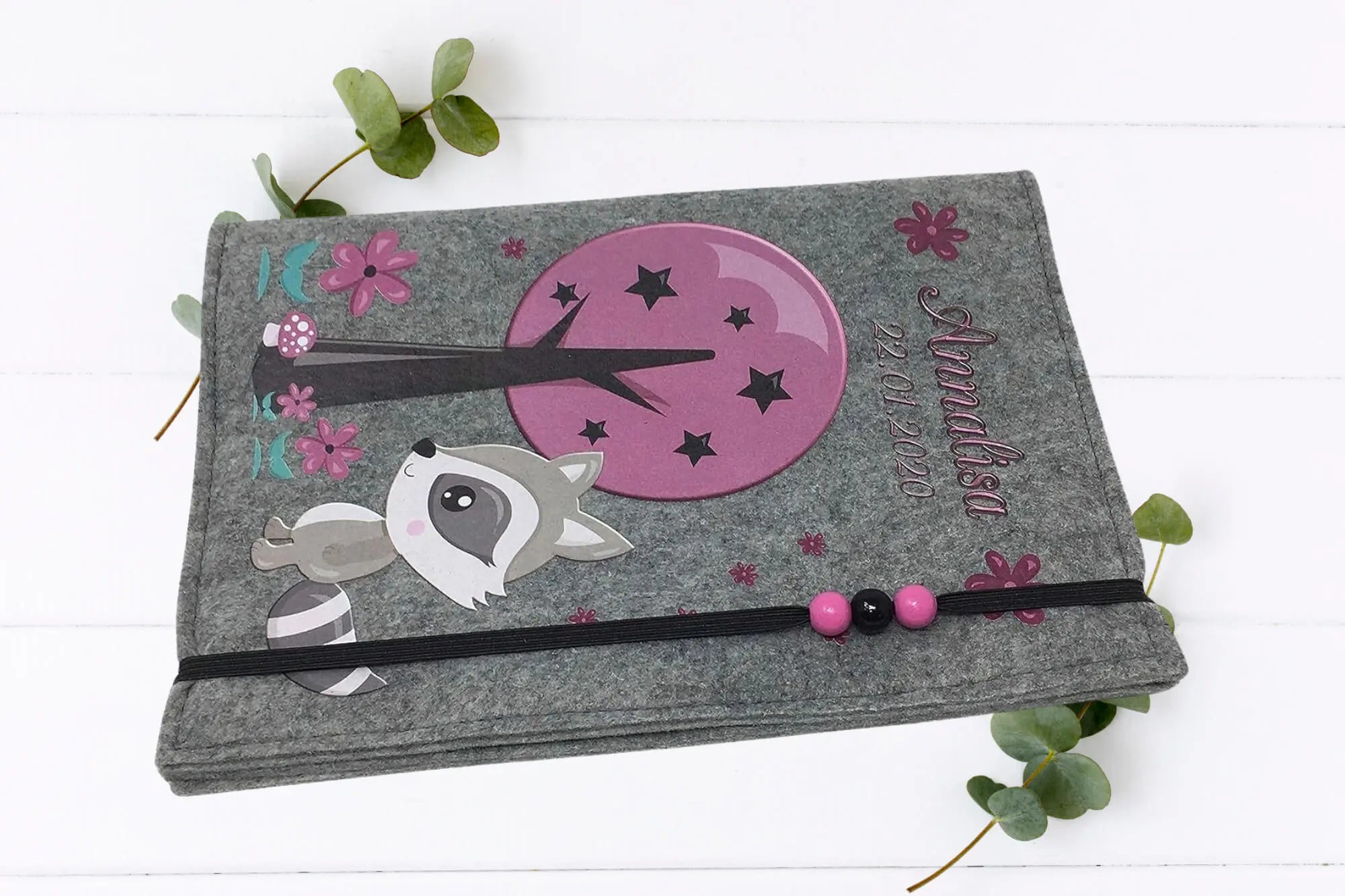 U-Heft-Hülle aus grauem Filz mit dekorativem Waschbär, lila Baumkrone und Blumenranke.