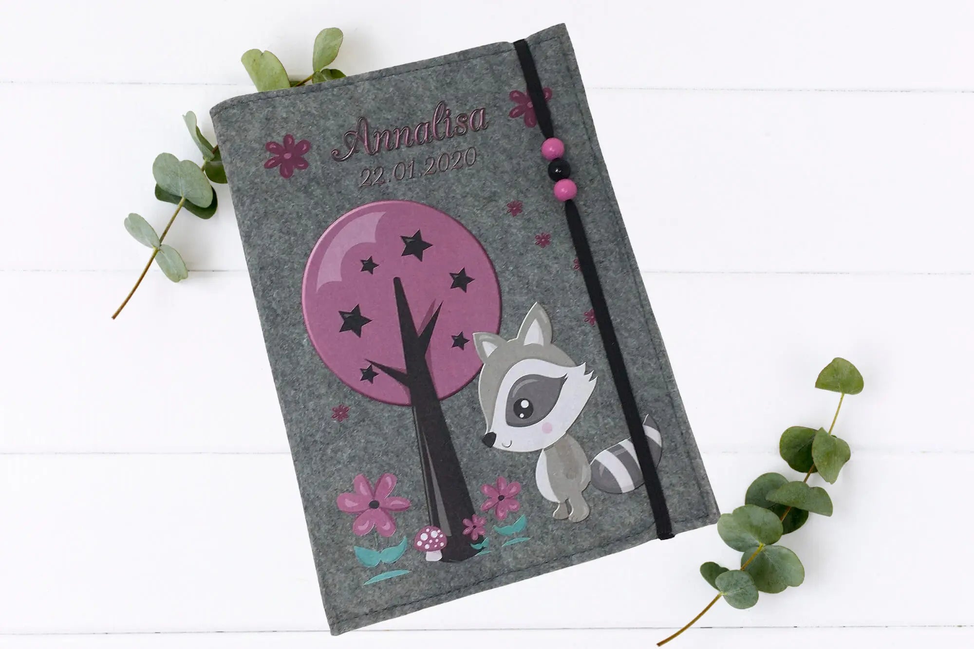 Filz-U-Heft-Hülle mit Waldmotiv: Waschbär in lila Landschaft mit Baum, Blüten und Perlenverschluss.