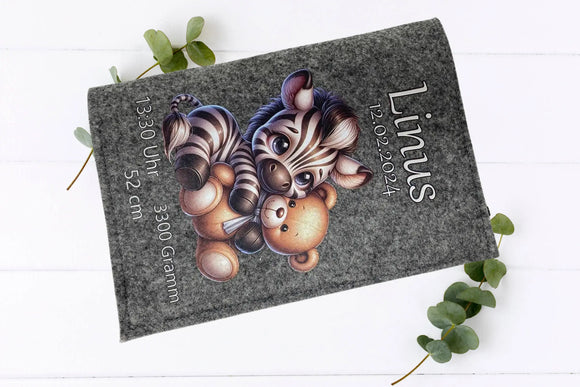Graue Filzhülle mit Zebra-Baby und Kuschelbär – emotionales Motiv für Babygeschenke.